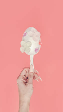Modern flat lay Easter concept. A female hand holds a white egg on a stick. Decoration with pastel...