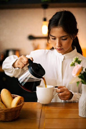Warm morning scene of a young woman pouring freshly brewed coffee into a white cup at a cozy kitchen table. Natural light, relaxed atmosphere, and breakfast setting with bread and flowers create a homely and peaceful mood.の素材