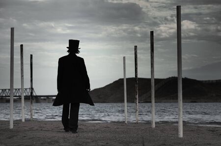 Rear view on alone man in the black coat and top-hat going to the sea. Natural darkness. Artistic...