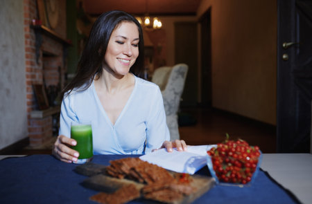 Smiling vegetarian woman sitting at the table with celery fresh juice, flaxseed bread and berries and reading bookの写真素材