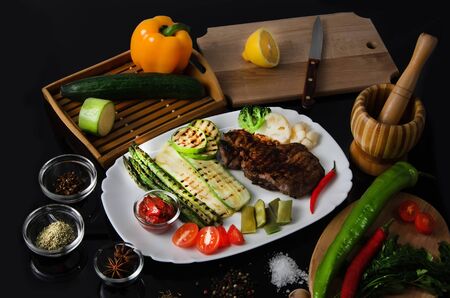 Grilled beef grilled steak with asparagus, zucchini, lobi, broccoli, tomato, chili and red sauce on a white plate, on a black glossy backgroundの写真素材