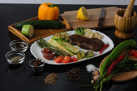 Grilled beef grilled steak with asparagus, zucchini, lobi, broccoli, tomato, chili and red sauce on a white plate, on a black glossy backgroundの写真素材