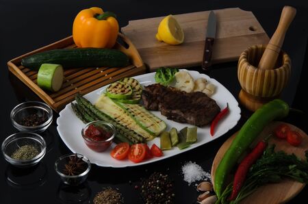 Grilled beef grilled steak with asparagus, zucchini, lobi, broccoli, tomato, chili and red sauce on a white plate, on a black glossy backgroundの写真素材