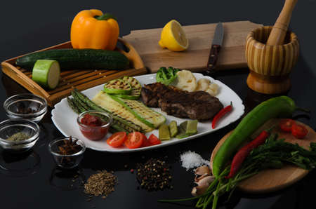 Grilled beef grilled steak with asparagus, zucchini, lobi, broccoli, tomato, chili and red sauce on a white plate, on a black glossy backgroundの写真素材