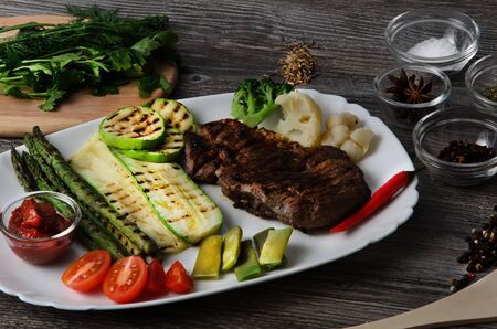 Grilled beef grilled with asparagus, zucchini, lobi, broccoli, tomato, chili and red sauce on a white plate, and other vegetables in wood backgroundの写真素材