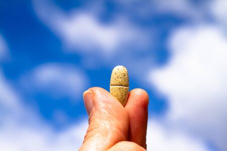 Vitamin pill held between fingers and thumb in a hand before a blue sky with white cloudsの写真素材