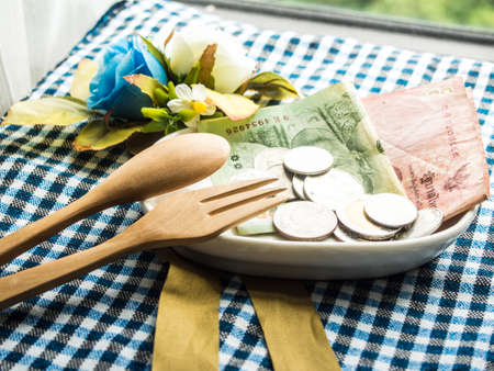 Thai baht note bank and coins in the white plate with wooden spoon and fork on the table