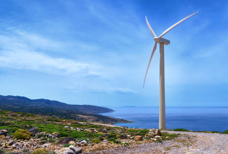 Single windmill turbine on hilltop of seashore in colorful landscape against dynamic blue sky with clouds and winding road. Crete, Greece.の写真素材