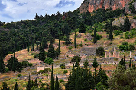 View of famous ancient Greek complex - Delphi oracle. Ruins of temple of Apollo and theater. Tourists rush up and down on paths among pines and cedarsの写真素材