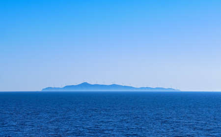 Seascape of calm azure sea, blue sky, no clouds. Distant island in haze with silhouettes of electric turbine windmills. Mediterranean sea, Greece.の写真素材