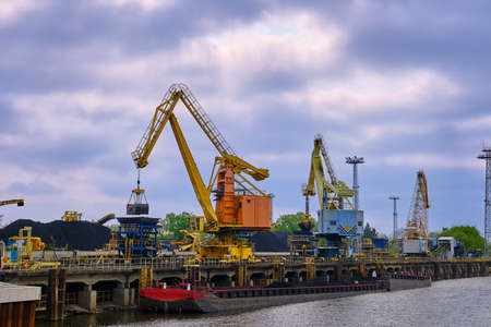 River port cranes with clamshell or griper loading coal to river drag boats or barges moored by pier on cloudy day. Energy, minerals, environmentの写真素材