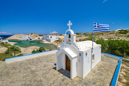 White Greek Orthodox chapel or church, white cross and Greek national flag on hilltop on sunny summer day. Greek island landscape. Milos, Greeceの写真素材