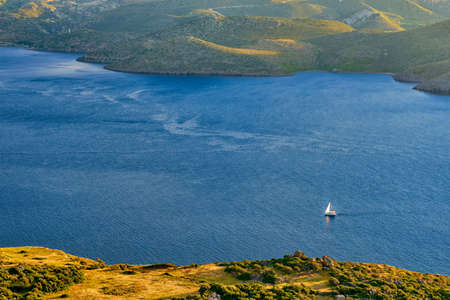 Lonely sailboat or yacht navigating in Aegean sea near Milos island on sunset. Distant hills, high coast, low sun, evening, Milos island, Greeceの写真素材