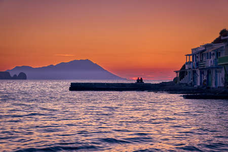 Sunset in fishermen village of Klima, Milos island, Greece. Pair of adults enjoy sunset view on pier of small fishermens village. Hazy hills, dawn.の写真素材