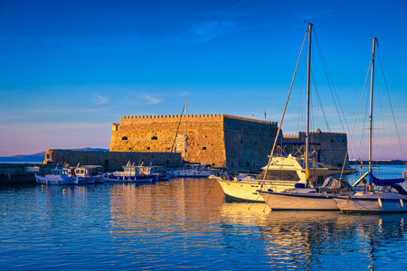 Landscape of Venetian castle or Koules fort in Heraklion, capital of Crete island, Greece and moored fishing boats at sunset. Clear blue sky, deep blue waters of port, yachts, sailboats. Travel destination of Mediterranean sea, summer vacations.の写真素材