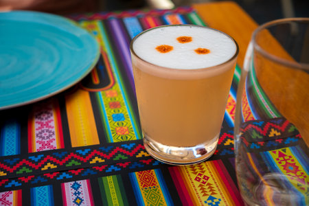 Pisco Sour alcoholic drink, Peruvian cocktail, made of Pisco (Peruvian grape schnaps), lime juice, syrup and egg white, in glass on placemats with traditional patterns. Selective focus, blurred background, focus on the front glass and foam.の写真素材