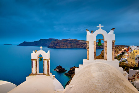 Two bell towers and dome roofs of white Greek Orthodox church in Oia village, Santorini island, against waters of Aegean sea, Greece, and distant island on overcast day with heavy clouds and dramatic sky. Famous local landmark, wedding destinationの写真素材