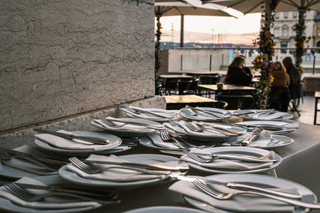 Plates neatly arranged with forks, knives, and napkins on stone countertop at outdoor restaurant in selective focus. Blurred background with European city square at sunset. Relaxed dining, people seated, umbrellas, travel destination, summer vacationの写真素材