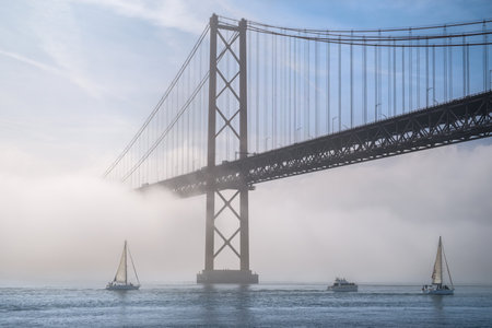 Dramatic view of Lisbon iconic 25 de Abril bridge in morning fog on sunny day, with sailboats and motorboat on tranquil Tagus river, Portugal. Misty atmosphere, serene, mysterious scene, perfect for travel, cityscape themes, suspension bridge.の写真素材