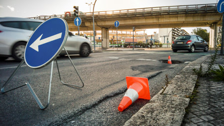 City street with large pothole marked by traffic cones and detour sign, cars passing by and overpass in background. Urban infrastructure issues, road safety, busy traffic hours, street repairs, traffic disruption, asphalt melting, summerの写真素材