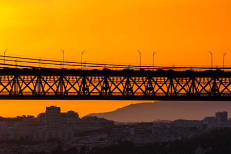 Bridge spans city skyline, silhouetted against vivid orange sunset. Soft city outlines and distant hills frame flowing late evening traffic, dynamic urban energy, tranquil evening light for unique harmony of architecture and nature. Urban landscapeの写真素材