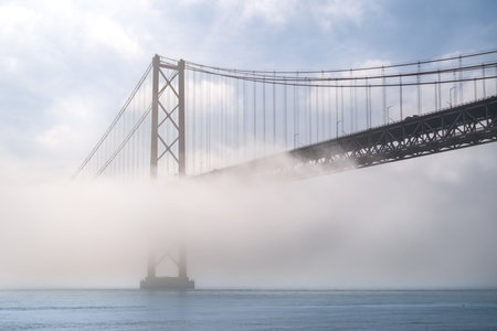 Iconic 25 de Abril suspension bridge, Lisbon, Portugal, in morning fog on sunny day over calm Tagus river waters. Misty atmosphere, peaceful, quiet scene, mist roll in over waters, riverscape, beauty in nature, tourist attraction, travel destinationの写真素材