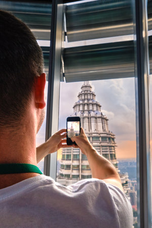 Nov 20, 2019 - Kuala Lumpur, Malaysia: Unrecognizable male adult tourist taking photo of Petronas tower with smartphone through window at colorful sunset.Observation deck visit, tourist attraction, travel destination, trip experience, famous landmarkの写真素材