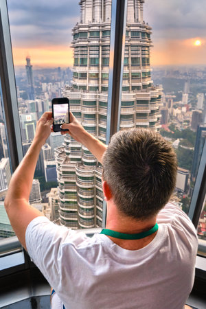 Nov 20, 2019 - Kuala Lumpur, Malaysia: Unrecognizable male adult tourist taking photo of Petronas tower with smartphone through window at colorful sunset.Observation deck visit, tourist attraction, travel destination, trip experience, famous landmarkの写真素材