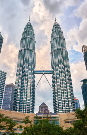 Nov 20, 2019 - Kuala Lumpur, Malaysia: Petronas Twin Towers with skybridge in urban skyline at day under dramatic cloudy blue sky. Scenic cityscape with lush park, modern Suria KLCC shopping mall in foreground, famous landmark, high-rise buildingsの写真素材