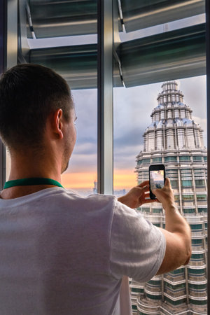 Nov 20, 2019 - Kuala Lumpur, Malaysia: Unrecognizable male adult tourist taking photo of Petronas tower with smartphone through window at colorful sunset.Observation deck visit, tourist attraction, travel destination, trip experience, famous landmarkの写真素材
