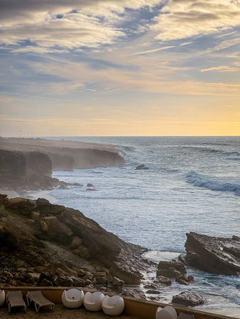 Scenic view of turbulent ocean waves hitting rugged cliffs as sun sets in background. White lounge chairs are scattered on terrace balcony, serene atmosphere. Ocean view, sundown sky with clouds.の写真素材