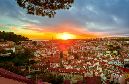 Beautiful sunset over Lisbon, Portugal, cloudy sky, sun setting behind April 25th or 25 de Abril bridge, over rooftops, Tagus or Tejo river, Sao Jorge castle hill. Travel destination, tourist attraction, sundown landscape, cityscape, trees, landmarksの写真素材