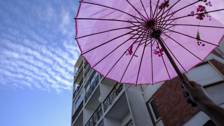 Feb 10, 2024 - Lisbon, Portugal: upshot of human hand holding pink Asian-style paper umbrella in selective focus against blue sky and some building in blurred background on sunny day. Symbol of pink theme of Colombina Clandestina carnival paradeのeditorial素材