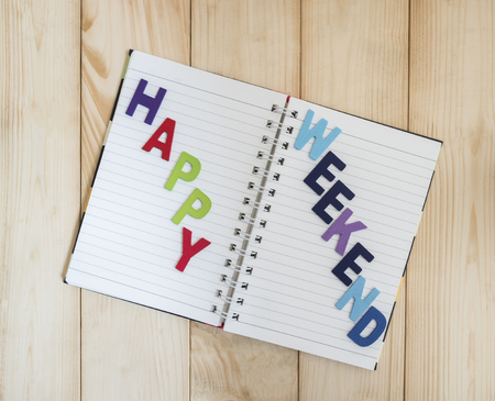 Word spelling Happy Weekend on notebook page with wood background (Business concept)