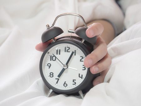 Close up hold alarm clock on white bed in the morning by woman hand with soft tone color.