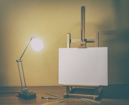 easel with canvas and brushes with a lit lamp on wooden tableの写真素材