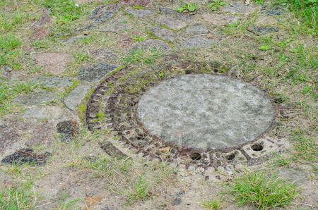 old manhole sewer cap in a brick pavement overgrowing with grass