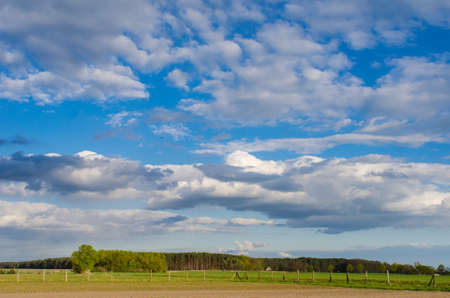 amazing cloudy sky over a rural landscape with forest and wooden fence on a sunny spring dayの写真素材