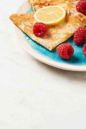 Folded pancakes with lemon slices and raspberries on white marble background