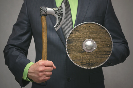 Businessman holds in hand a toy viking axe weapon and shield. Life or finance insurance agent. Money deposite safety agent.の写真素材