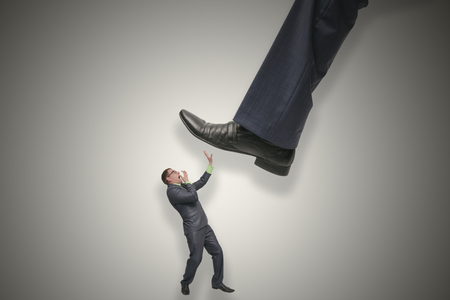 A huge leg of a man steps on a small frightened weak businessman isolated on gray background. Angry arrogant boss. Walk on the heads concept.の写真素材