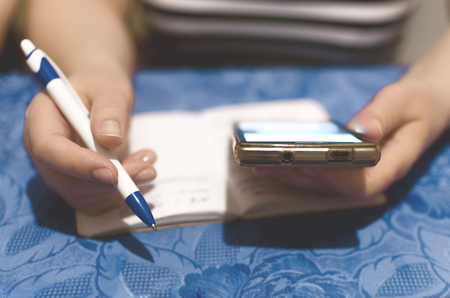 Woman is writing a notes in a notebook, plans daily schedule and is looking on her mobile phone in her hand.の写真素材