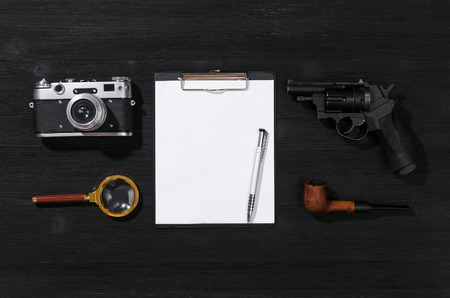 Blank page, pen, film photo camera, magnifying glass, handgun and smoking pipe on a black detective agent table background. Secret document mockup. Detective agent report. Flat lay spy table concept.の写真素材