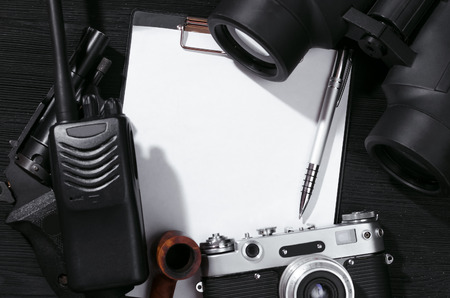 Blank page, portable radio, handgun, binoculars and photo camera on a black flatlay table...