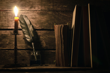 Quill pen in a inkpot and stack of books in the light of burning candle on the writer desk background. Education concept.の写真素材