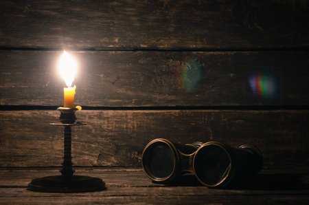 Binoculars and burning candle on the wooden table background with copy space. Travel or adventure concept.の写真素材