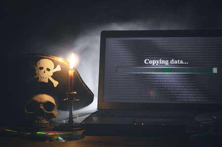Illegal data copying concept. Cybercrime. Computer piracy background. Pirate hat, human skull, laptop and compact disc on a table.の写真素材