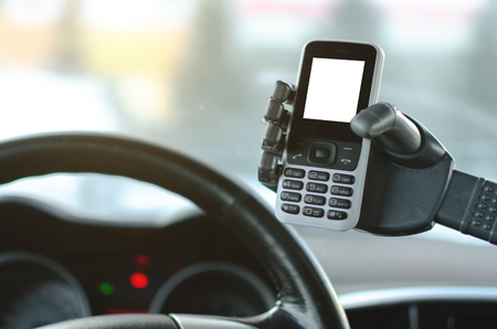 Mobile phone with blank screen in a robotic hand on a car steering wheel and dashboard background. Handsfree driving or automatic driver assistant concept.の写真素材