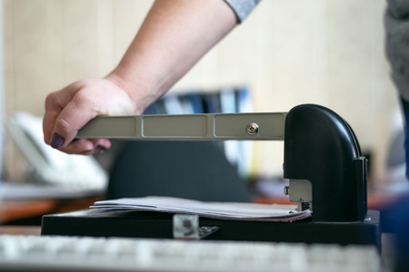 Office worker is punching a paper sheets with hole puncher. Paperwork background.