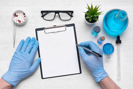 Medical examination mockup. A doctor or pharmacist is writing a notes to a blank paper sheet with a pen in his hand on laboratory table background.